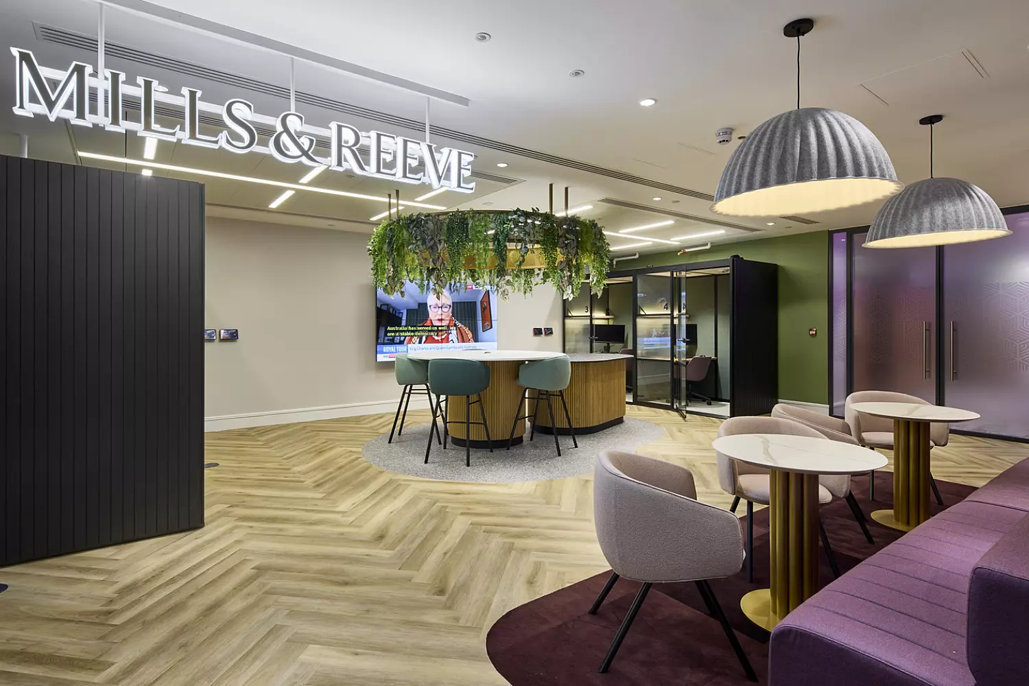 A contemporary office interior with the name "MILLS & REEVE" displayed on a ceiling-mounted sign. The space includes a variety of seating options, such as round tables with chairs and a long purple bench along the wall. A large central table is positioned beneath hanging plants, adding a natural touch. The flooring features a patterned wood design, while the walls are painted in soft purple and beige tones, accented with decorative wooden panels. Lighting is provided by a mix of recessed fixtures and pendant lamps.