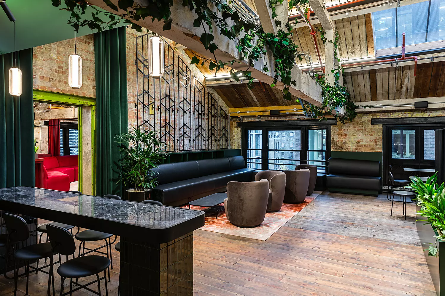A stylish office lounge with an industrial chic design, featuring exposed brick walls, wooden flooring and large skylights that let in natural light. The space is furnished with black upholstered bench seating, plush brown armchairs and a dark marble high top table with matching black stools. Greenery, including hanging plants and potted plants, adds a refreshing touch. Decorative metal screens and a green velvet curtain frame an entrance to another room, where red seating is visible. The overall atmosphere is a blend of modern sophistication and rustic charm.