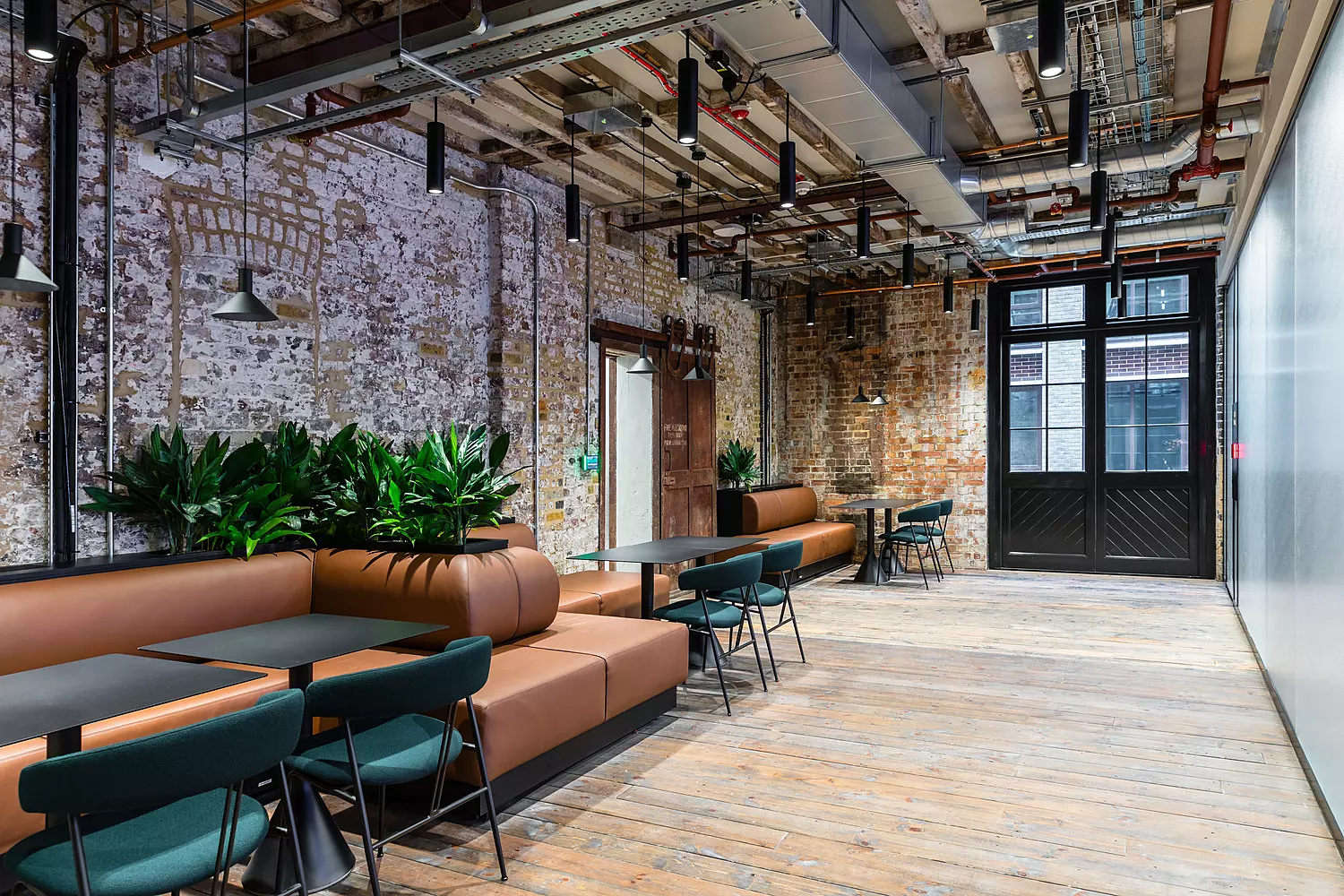 A modern industrial-style office space featuring exposed brick walls, wooden flooring, and an open ceiling with visible pipes and ductwork. The room is furnished with long brown leather banquettes paired with dark green chairs and black tables, creating a stylish seating area. Pendant lights hang from the ceiling, adding to the urban aesthetic. Green plants placed along the banquettes provide a refreshing contrast. At the far end, large black-framed double doors lead to another area, allowing natural light to filter into the space.