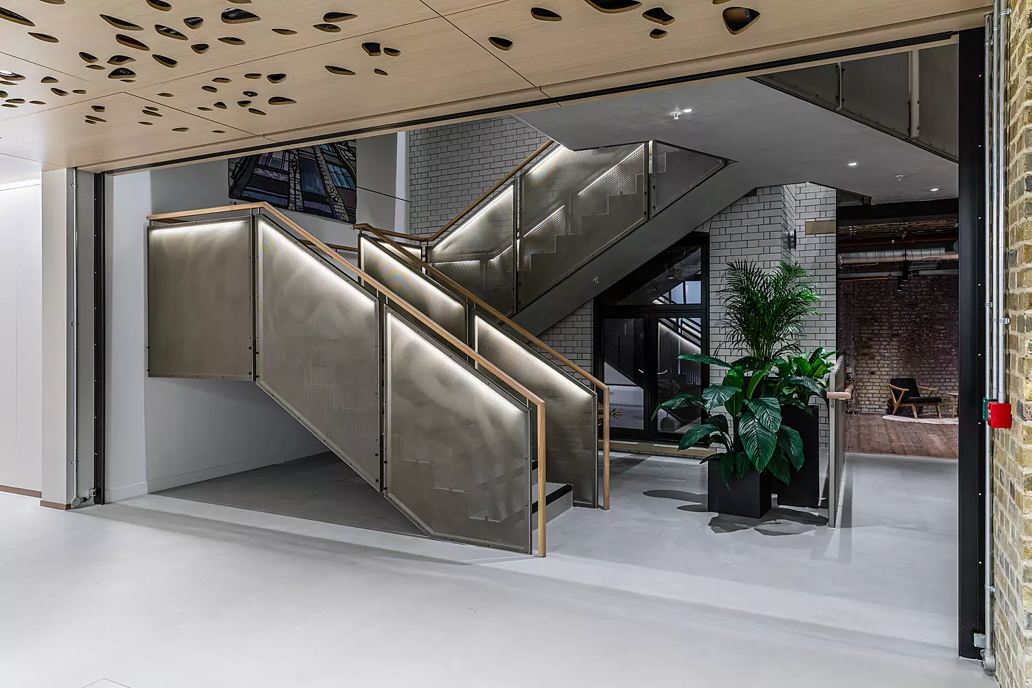 A modern office interior featuring a striking metal staircase with illuminated handrails. The space has an industrial aesthetic with exposed brick walls, white subway tiles and polished concrete flooring. Green plants in sleek black planters add a touch of nature. The ceiling showcases a perforated wooden panel design, and in the background, a seating area with lounge chairs is visible, creating a stylish yet functional workspace.