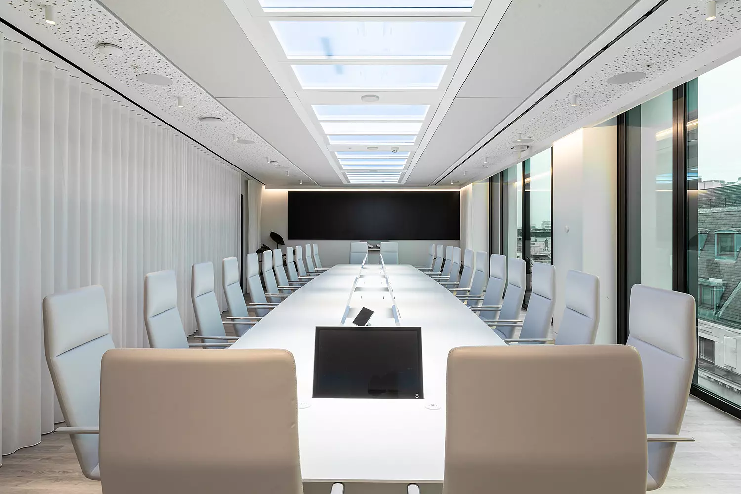 A sleek, modern boardroom with a long white conference table surrounded by high backed white chairs. The room is illuminated by skylights and floor-to-ceiling windows on one side, offering a view of surrounding buildings. A black screen is mounted on the far wall, and built-in screens are embedded in the table for digital collaboration. The ceiling features perforated acoustic panels, and a white curtain runs along one wall, adding a soft contrast to the clean, minimalist design.
