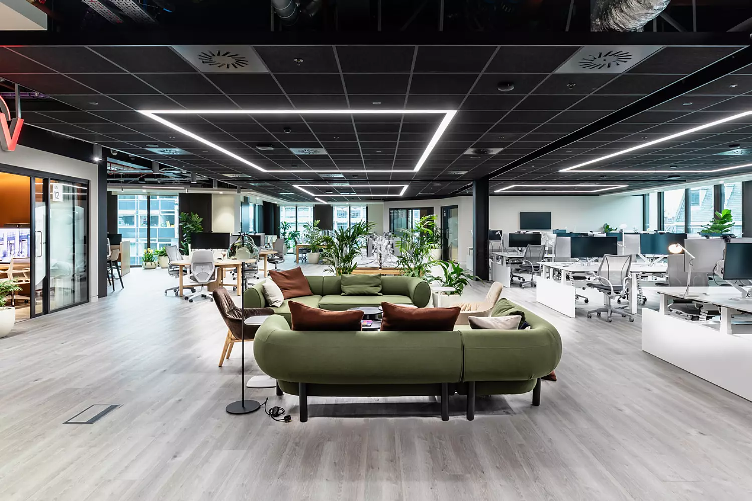 The space features white desks with ergonomic chairs, large monitors, and a central lounge area with green sofas and brown cushions. There are several potted plants throughout, and the ceiling has black panels with integrated LED lighting. A glass meeting room is visible on the left with the "ViiV" logo above it. The office has large windows allowing natural light to enter.