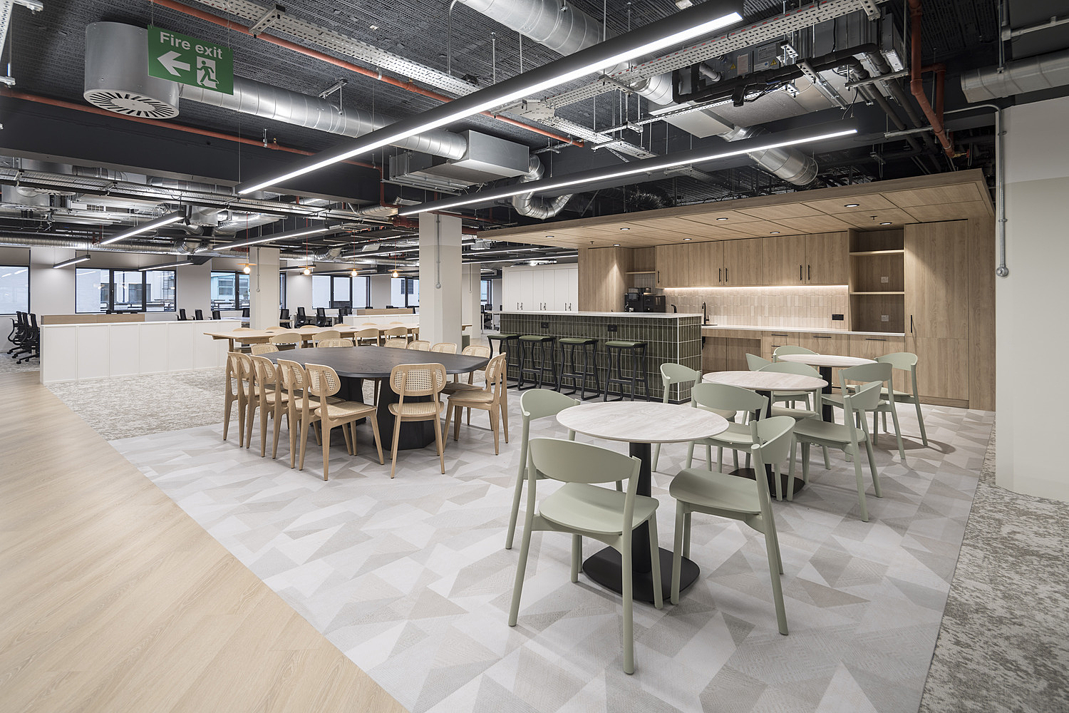 modern open-plan office space featuring a mix of round and rectangular tables with chairs arranged in the foreground. In the background, there is a kitchen or bar area with stools lined up along a counter. The ceiling has exposed pipes and ductwork, contributing to an industrial aesthetic. An emergency exit sign is visible on the ceiling towards the left side of the image.