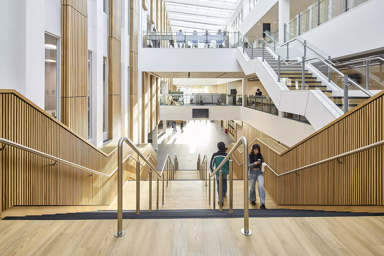 A spacious, modern interior featuring wide wooden staircases with sleek metal railings, descending to a lower level. The walls and railings are finished in warm-toned wood, complemented by large windows on the upper levels that flood the space with natural light. Glass barriers line the edges of the upper floors, enhancing the open and airy feel. A few people are visible on the stairs and upper levels, adding a sense of scale and activity.