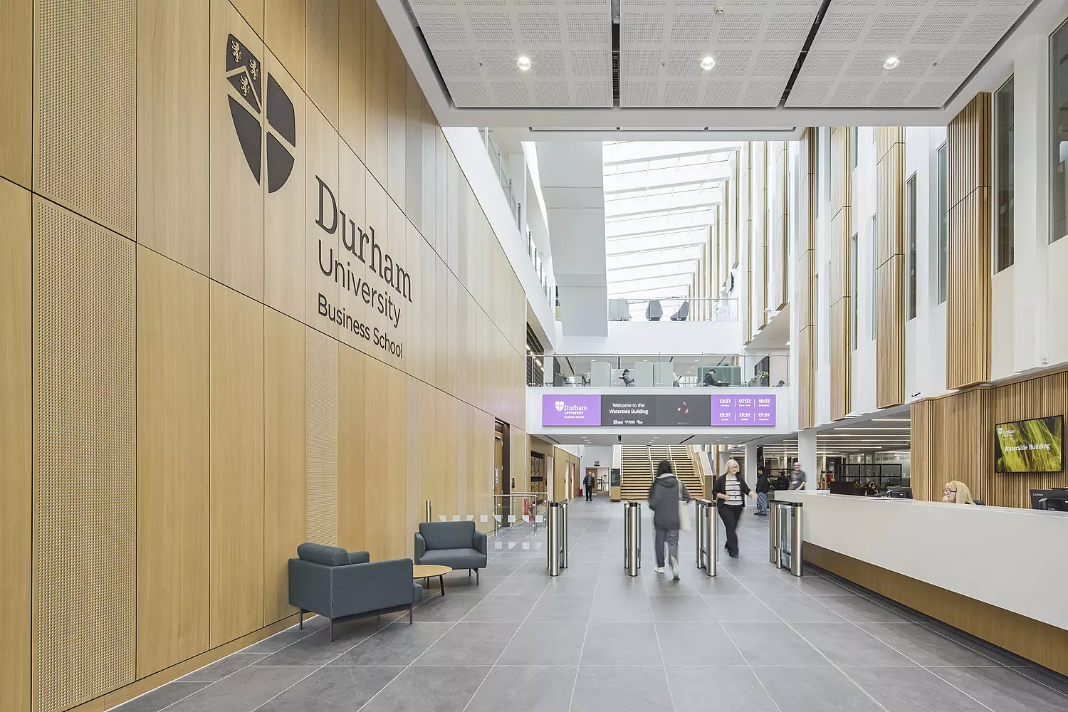 A contemporary interior space within Durham University Business School, featuring wooden panelled walls and a high ceiling with integrated lighting. Large windows allow ample natural light to flood the area. The space includes modern seating arrangements with chairs and sofas, an information desk on the right, and turnstiles near the entrance. The university’s logo and name are displayed prominently on the left wall.