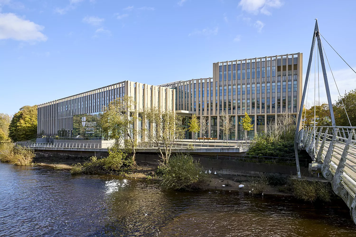 A contemporary building with a façade of large glass windows and vertical wooden slats, positioned beside a tranquil body of water. Trees and vegetation line the water’s edge, enhancing the natural setting. A pedestrian bridge extends from the right, leading towards the building. The sky above is mostly clear with a few scattered clouds, contributing to the serene atmosphere.