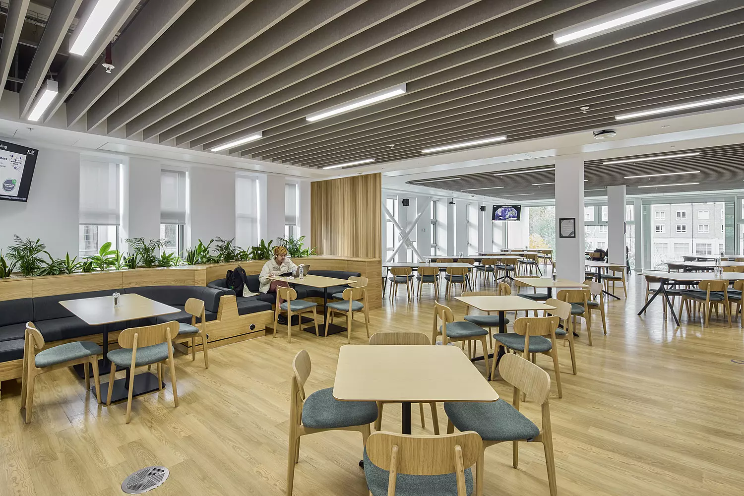 A modern, spacious cafeteria dining area with wooden flooring and matching wooden tables and chairs arranged in an orderly layout. The ceiling features parallel beams with recessed lighting, adding a clean, structured aesthetic. Large windows line one side of the room, allowing natural light to brighten the space. Potted plants along the seating area introduce a touch of greenery, and a few people are seated at various tables.