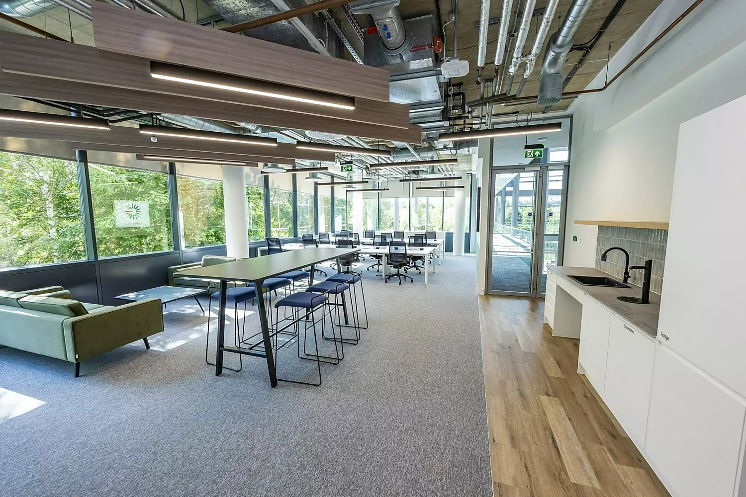 A modern, open-plan office space featuring two green sofas, a seating area with blue barstools, and a stylish green-tiled section. Rows of white desks with black ergonomic chairs provide a functional workspace, while feature lighting adds warmth and character. The design blends contemporary and industrial elements, creating a balanced mix of comfort and productivity.
