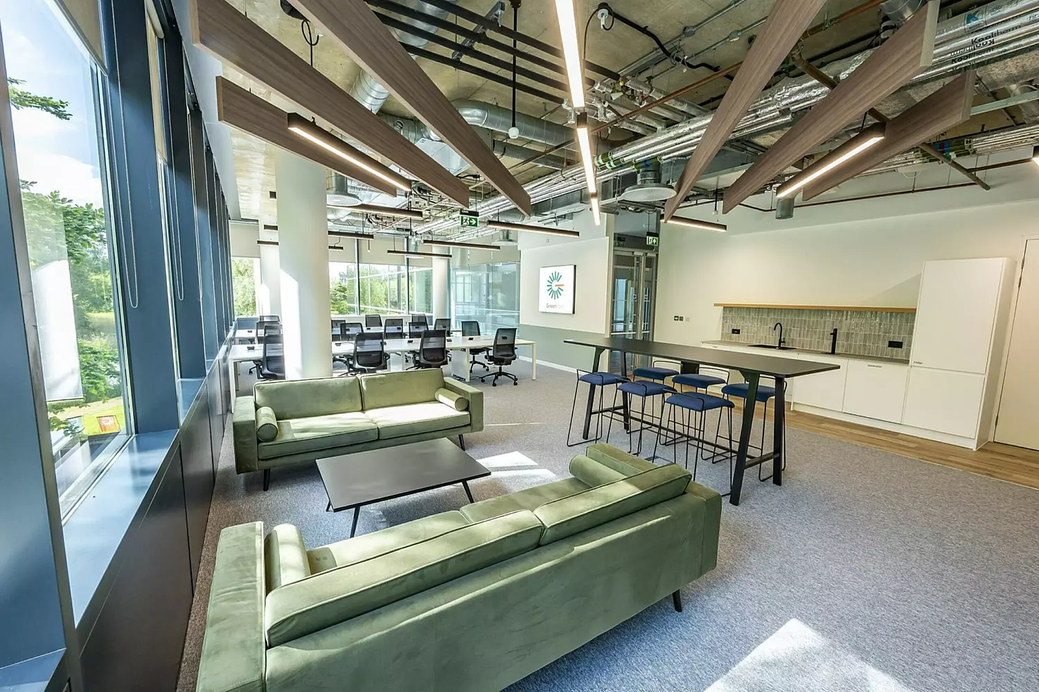 A modern, open-plan office space featuring two green sofas, a seating area with blue barstools, and a stylish green-tiled section. Rows of white desks with black ergonomic chairs provide a functional workspace, while feature lighting adds warmth and character. The design blends contemporary and industrial elements, creating a balanced mix of comfort and productivity.