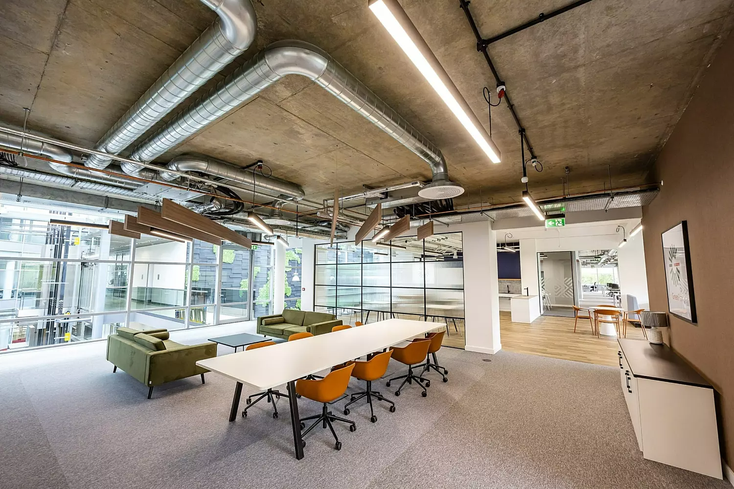 A modern office space featuring two green sofas and stylish feature lighting. A large white table is surrounded by vibrant orange chairs, creating a collaborative workspace. A frosted glass partition at the back adds a sense of privacy while maintaining an open feel. The design blends bold colours with contemporary elements for a dynamic and inviting environment.