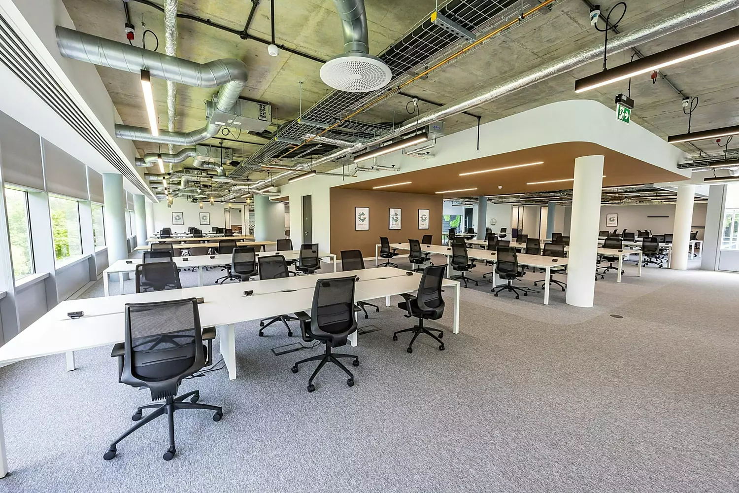 A spacious, modern open-plan office with rows of white desks and black ergonomic chairs. The office has an industrial-style ceiling with exposed ductwork, suspended lighting, and cable trays. Large windows along one side let in natural light, while neutral grey carpeting covers the floor. The space is divided by a partial brown partition with framed artwork. The overall design is clean and minimalistic, with a focus on functionality and flexibility.