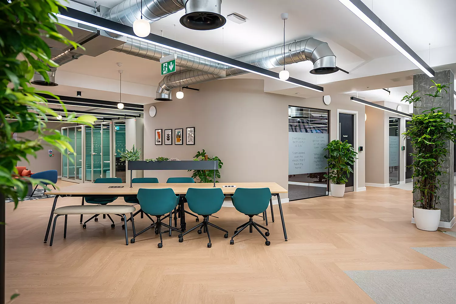 Modern office workspace with a large shared wooden desk surrounded by teal swivel chairs. The area has light wood flooring, potted plants and exposed ductwork on the ceiling with hanging lights. There are framed prints on the curved wall in the background, and glass-walled meeting rooms are visible to the left and right. The design is open and airy, with a clean and professional feel.