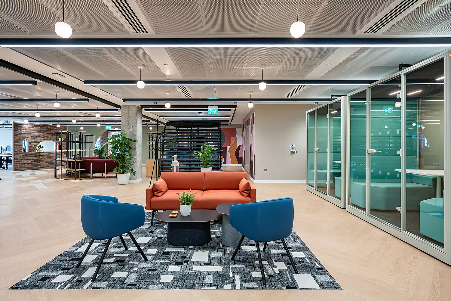 Modern office lounge area with a bright orange sofa, two blue armchairs and a geometric black and white rug. The space features light wood flooring, glass meeting rooms on the right, and an open-plan layout with bookshelves, plants and varied seating in the background. The ceiling has hanging lights and exposed panels, giving the room a contemporary feel.