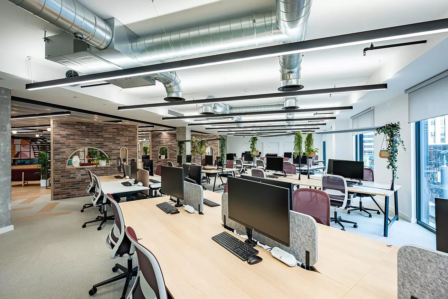 Open-plan office space with rows of light wooden desks, each equipped with desktop monitors, keyboards and chairs in burgundy and black. The ceiling features exposed ductwork and long black strip lights. Large windows on the right let in natural light, and there are several potted plants placed around the room. A brick feature wall with arched cut-outs adds a touch of character to the modern, airy setting.