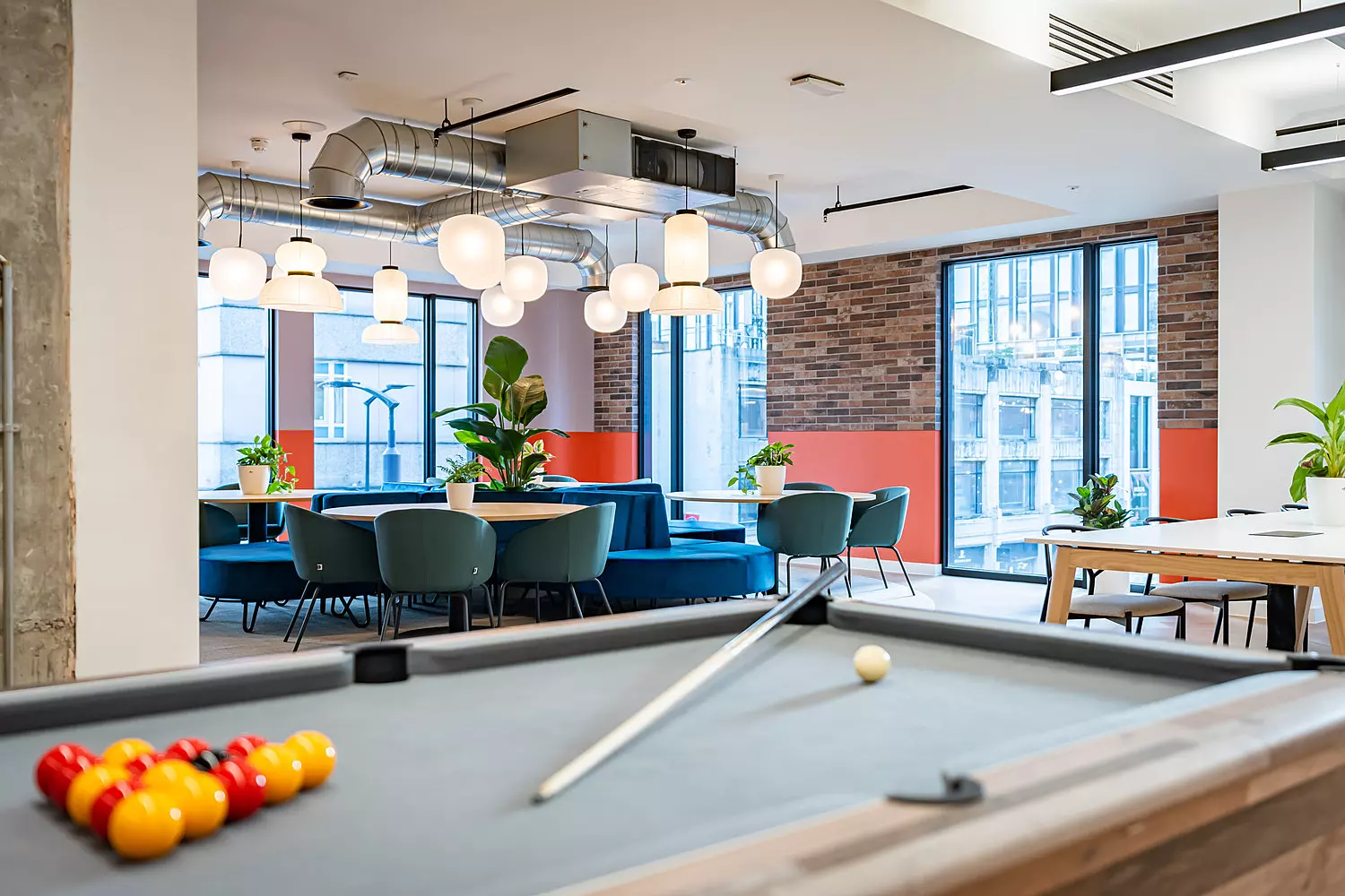 Social space in a modern office with a pool table and comfy seating. The walls have exposed brick and several full height windows letting through lots of natural light.