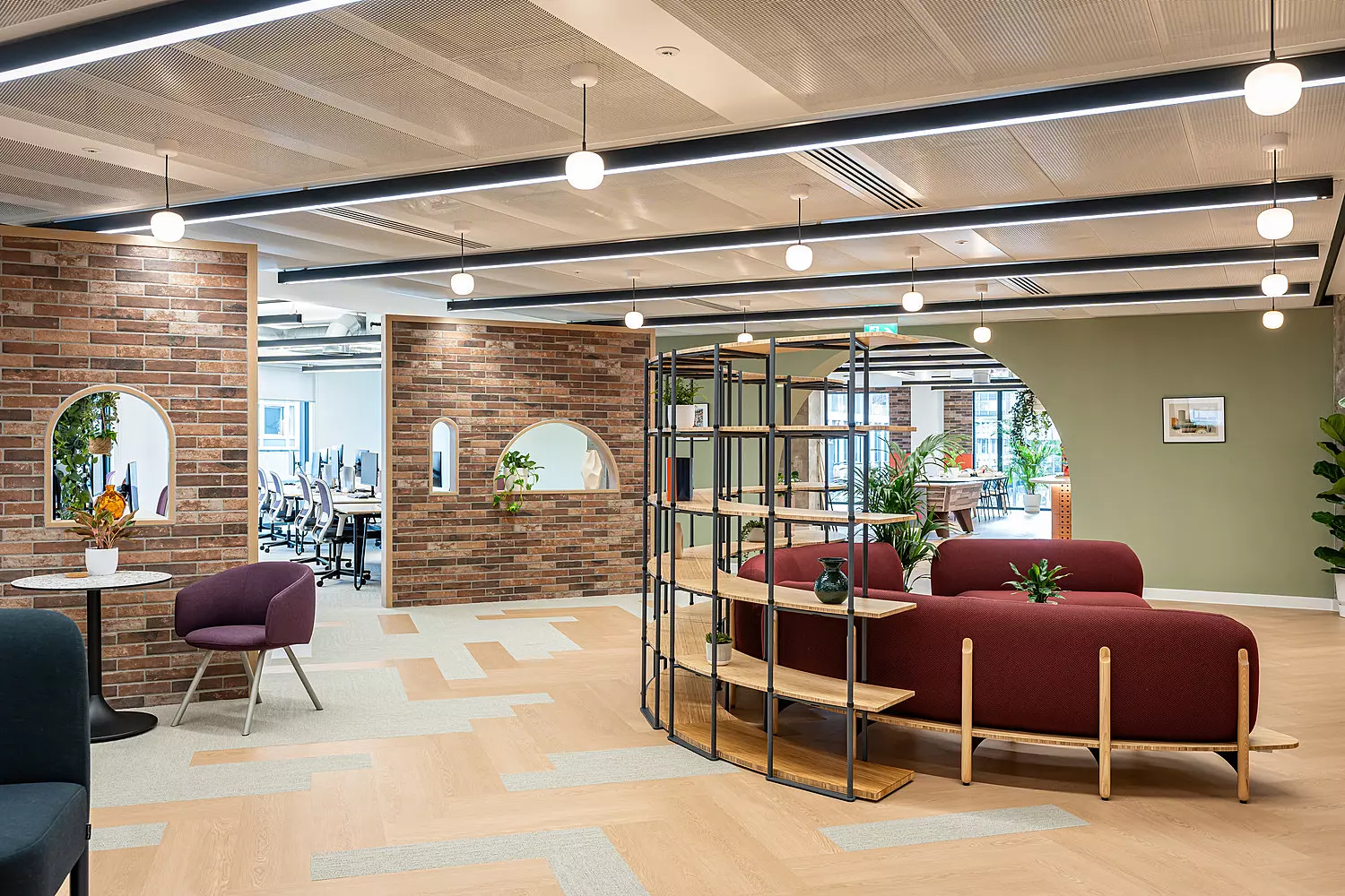 An open plan area of a modern office with warm exposed brick walls and wooden floors. There's a large semi-circular burgundy sofa with shelves behind it, as well as smaller chairs at small round tables.
