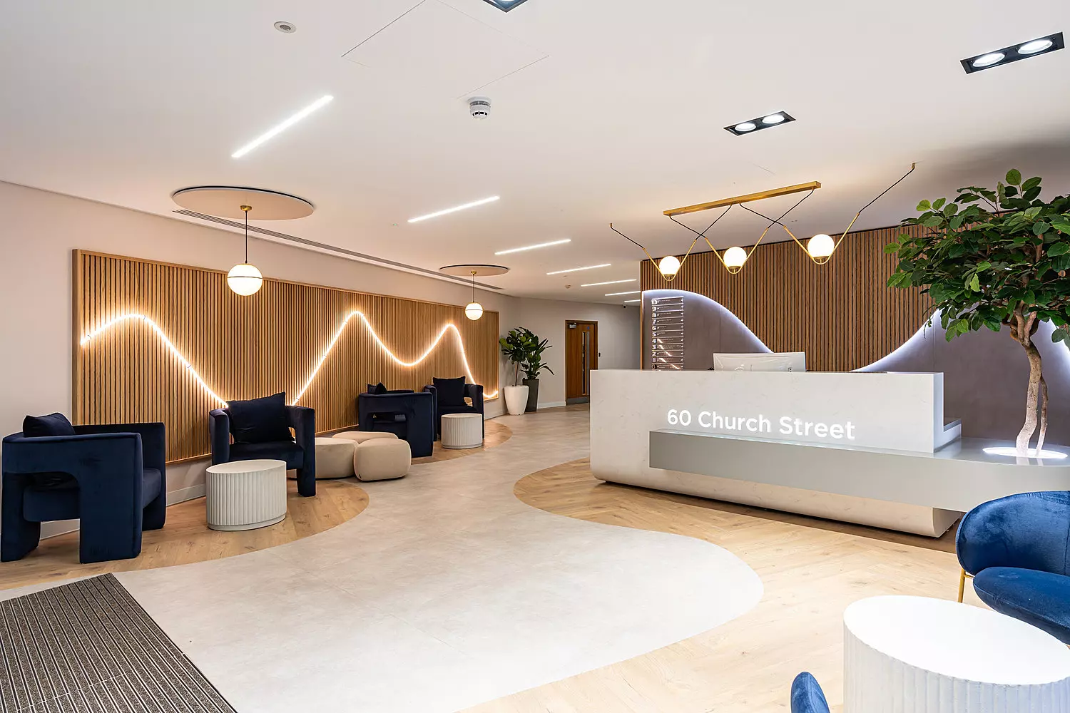A modern office reception area with a clean, minimalist design. The space features a light wood and white colour scheme with a wave-like LED light pattern on a slatted wooden wall. The reception desk is white with "60 Church Street" lit up on the front. There are navy blue armchairs, round pouffes and small white tables arranged in seating areas. Large potted plants add greenery and contemporary pendant lights hang from the ceiling. The flooring blends light wood and white finishes in a flowing pattern.