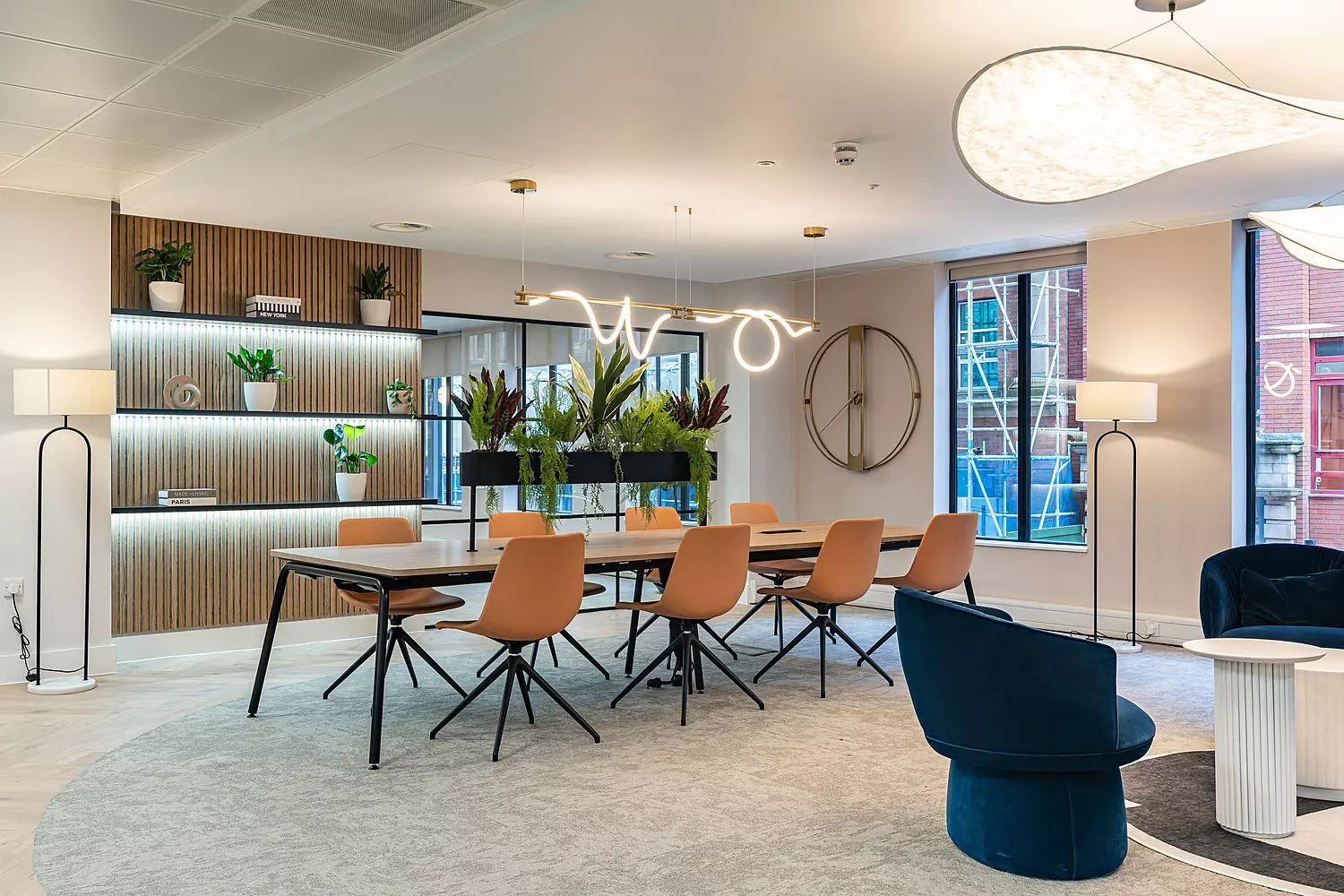 A stylish and open meeting area with a modern design. The room features a long wooden table surrounded by tan chairs with black legs, set on a light grey circular rug. A curved LED pendant light hangs above the table. Behind the table is a low row of potted plants acting as a natural divider. A wooden slatted wall with built-in shelving displays more plants and decorative items. Large windows bring in natural light, and the space is finished with contemporary floor lamps, dark blue lounge chairs and a soft, neutral colour palette.