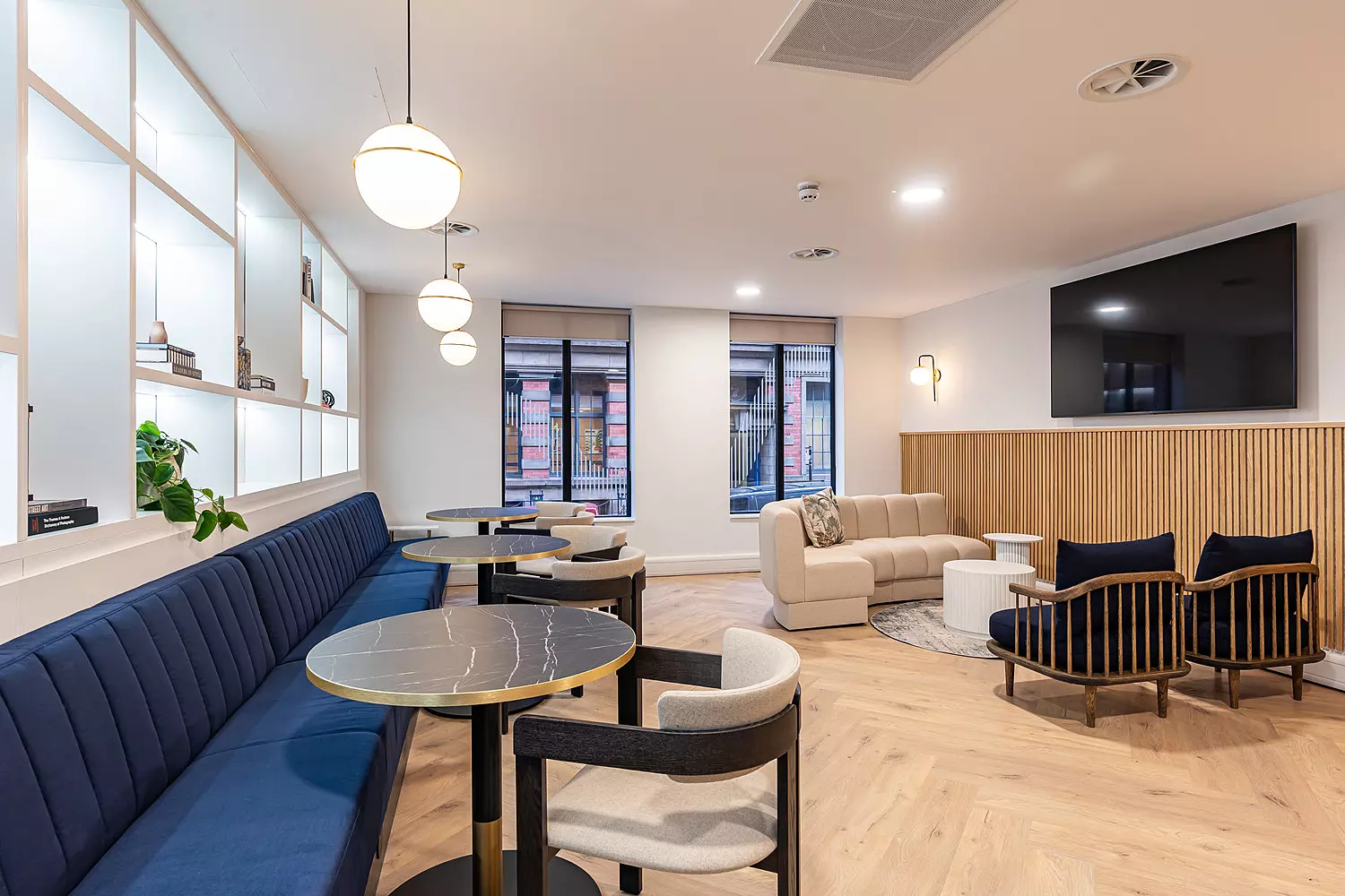 A relaxed breakout space with a mix of casual seating and modern finishes. The area includes a long navy blue bench with round black marble tables and cushioned chairs. There's a light beige sofa and two dark lounge chairs with wooden frames arranged around a small coffee table. The walls feature open shelving with plants and decor, and a large wall-mounted TV sits above wooden panelling. Pendant globe lights hang from the ceiling, and the room has light wood herringbone flooring and plenty of natural light from three large windows.
