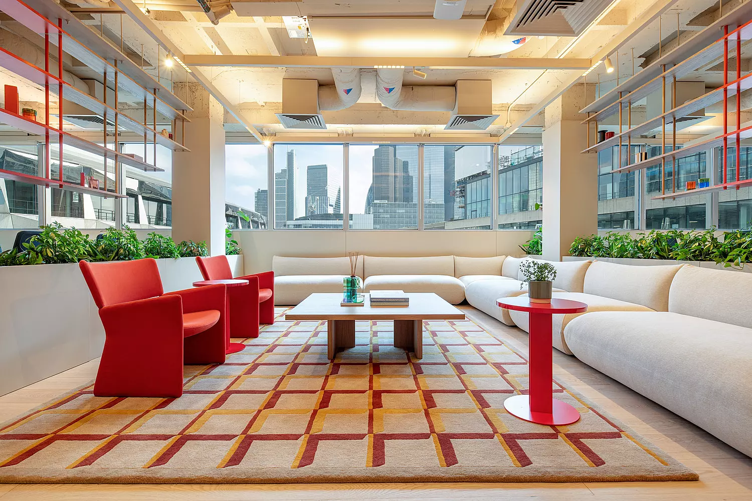 A bright office lounge area with large windows offering views of city buildings. The space includes a long white L-shaped sectional sofa arranged around a central coffee table, flanked by two red chairs and two small round tables, one red and one white. A patterned rug in beige and red covers the floor. Green plants line the edge of the seating area, and the ceiling features exposed ductwork and lighting fixtures.