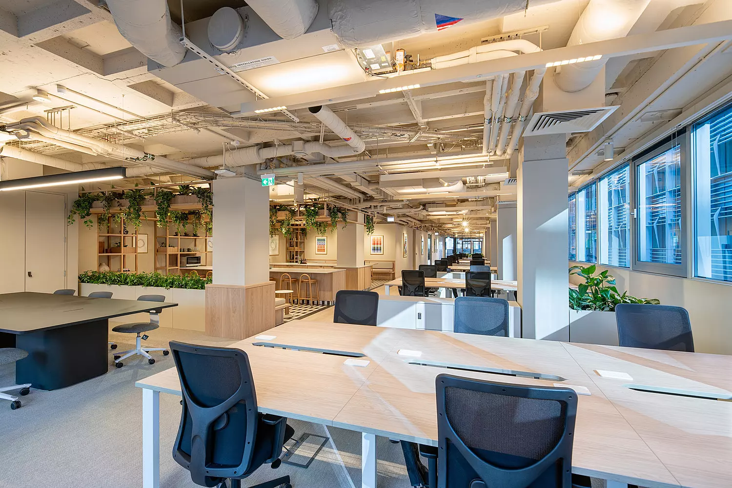 An open-plan office space with multiple workstations arranged in rows. Large windows on the right side let in natural light, while exposed pipes and ducts on the ceiling contribute to an industrial feel. The workspace includes desks and chairs throughout, with plants and greenery visible in the background adding a natural element to the environment.