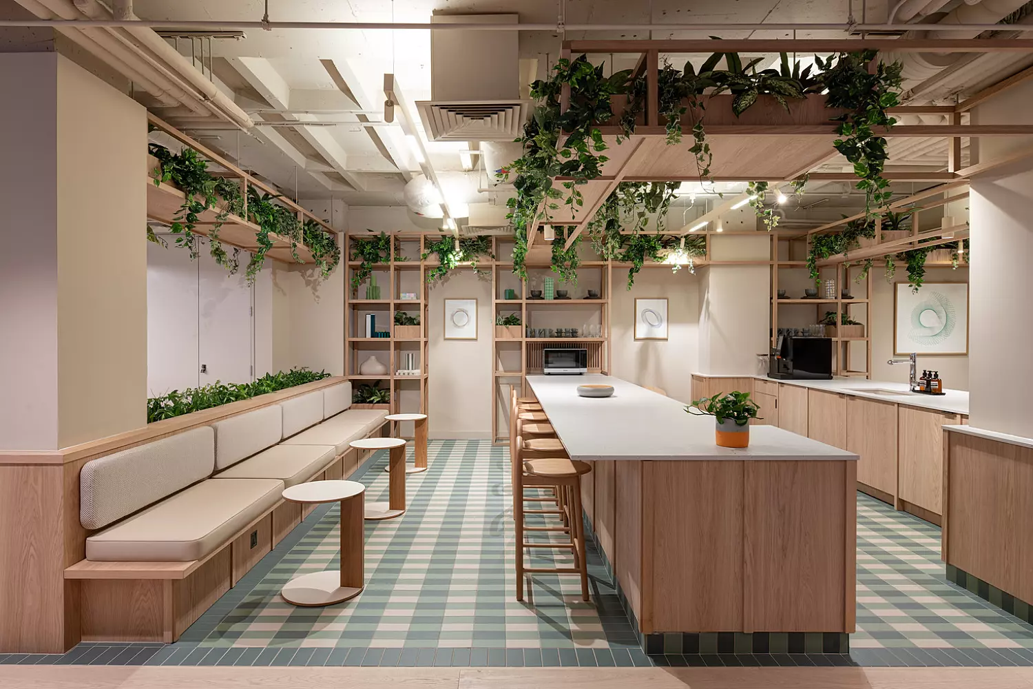 A bright teapoint area featuring wooden furniture and green plants throughout. Along the left side, cushioned benches are paired with small round tables. A large island counter with bar stools sits at the centre of the space, topped with hanging plants above. The floor has a green and white chequered pattern. Shelves on the walls hold books and decorative items, contributing to a clean, nature-inspired design.