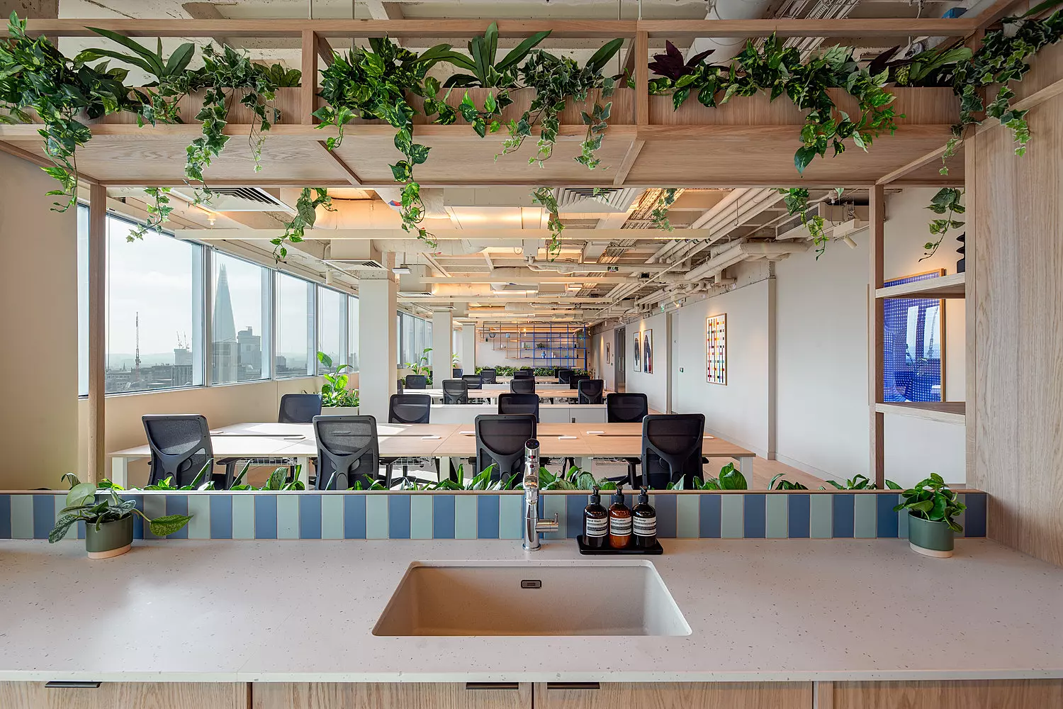 An open-plan office space with rows of desks and chairs arranged beneath a ceiling featuring exposed pipes and wooden beams. Large windows along one side allow natural light to fill the room. Hanging plants are suspended from the beams, adding greenery to the environment. In the foreground, a countertop with a sink and several bottles is visible. The design is clean and focused on natural materials and openness.