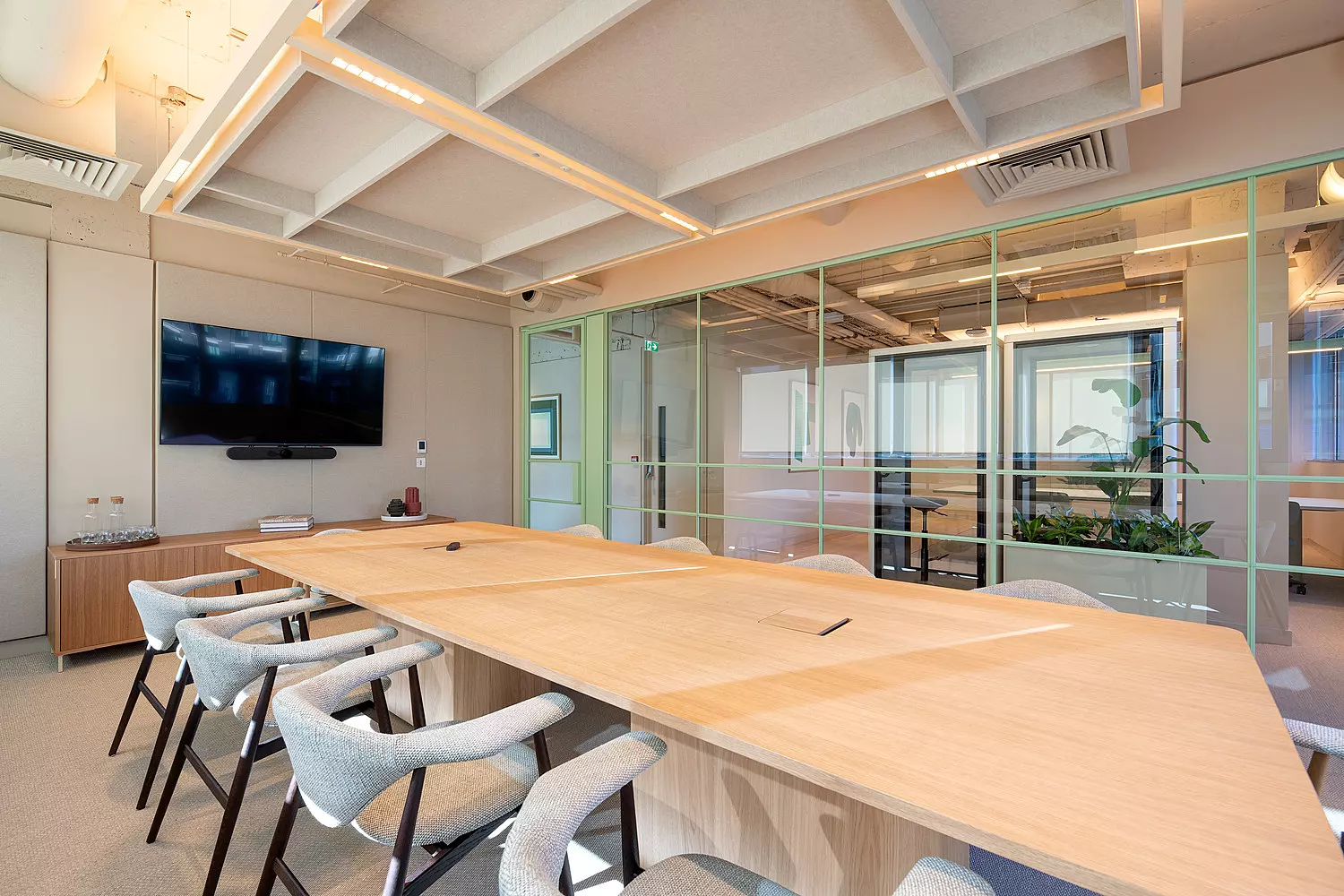 A conference room with a large wooden table surrounded by several chairs. The space is enclosed by glass walls, offering views into adjacent areas. A flat-screen television is mounted on one wall, and the ceiling is fitted with recessed lighting. The design is clean and streamlined, with a focus on transparency and functionality.