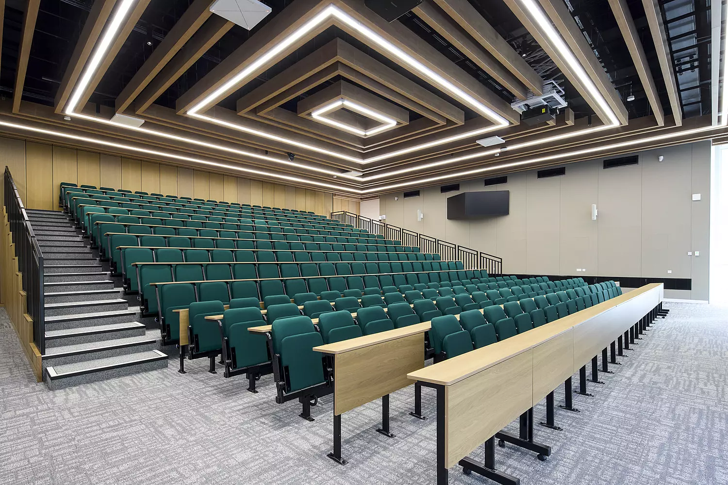 A modern lecture hall with tiered green seating arranged in rows, divided by a central aisle. At the front, a long desk spans the width of the room. The beige walls and contemporary ceiling lighting contribute to a clean, professional atmosphere. A large screen is mounted on one wall, enhancing the space’s functionality for presentations and lectures.
