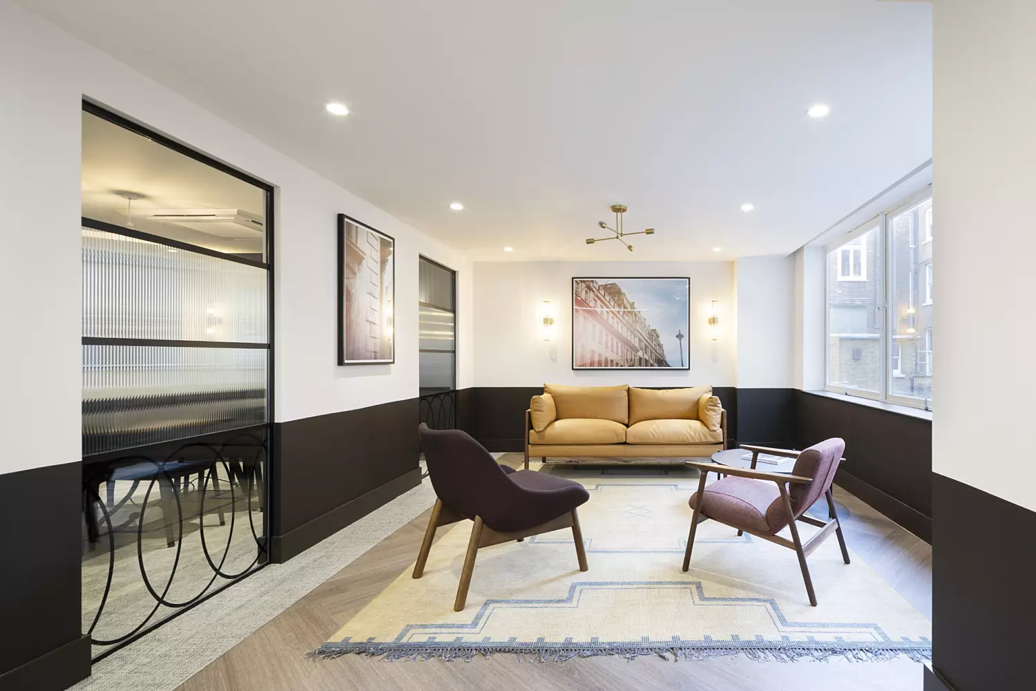 A bright, minimalist living room with a mustard-coloured sofa placed against the back wall, framed by two pieces of artwork. In front of the sofa is a light rug with geometric patterns, surrounded by a dark brown armchair and two wooden chairs with fabric seats. Large windows on the right side bring in natural light. The walls are painted white with black wainscoting, and a glass partition on the left separates the space from another area furnished with tables and chairs.