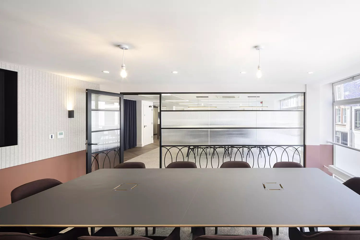 A well-appointed conference room with a large rectangular table surrounded by chairs. The space features white walls, with sections of brown panelling adding contrast. Two pendant light fixtures hang from the ceiling, and a large window on the right side allows natural light to brighten the room. The back wall is fitted with glass panels and a door that leads to an adjoining area.