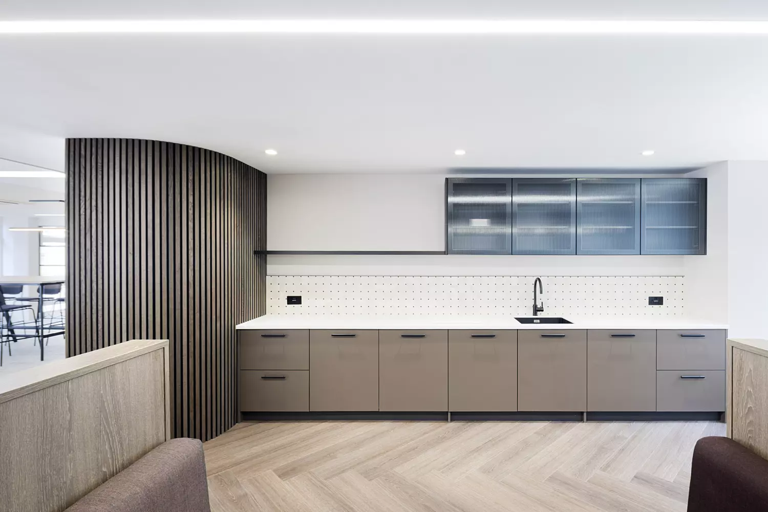 A sleek office teapoint with a minimalist design, featuring a long countertop with grey lower cabinets and glass-fronted upper cabinets. A black sink and faucet are centrally positioned along the counter, which is backed by white tiled splashback and illuminated by a strip of LED lighting beneath the upper units. On the left, a curved wall with vertical wooden slats adds a distinctive architectural detail. The floor is laid with light wood in a herringbone pattern.
