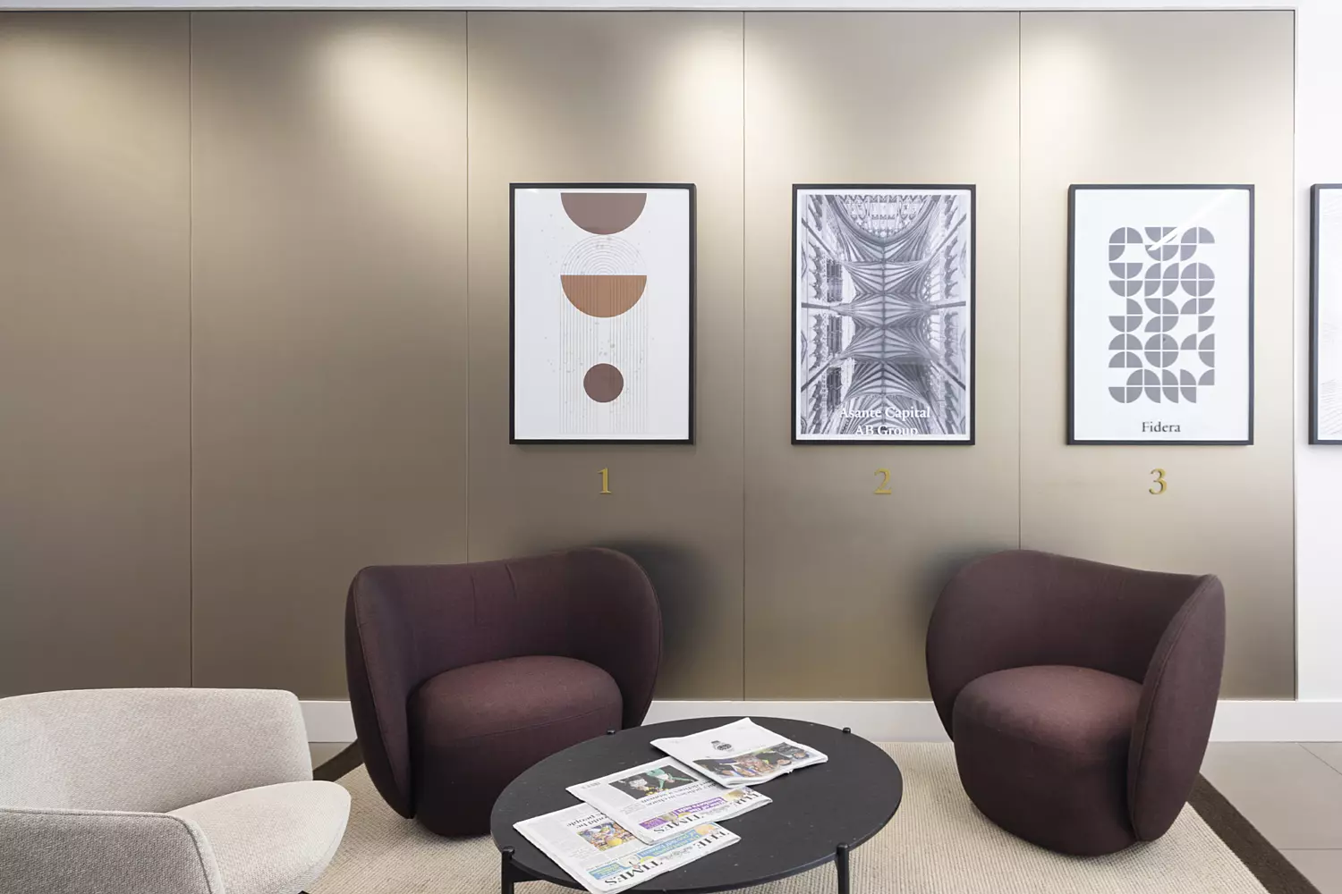 A minimalist waiting area or lounge furnished with two dark purple armchairs and one light grey armchair, arranged around a round black coffee table. Several magazines are placed on the table. The back wall features three framed artworks: one with circular abstract shapes, another resembling an architectural drawing, and a third displaying stylised text. The space is well-lit with ceiling lights, creating a clean and inviting atmosphere.