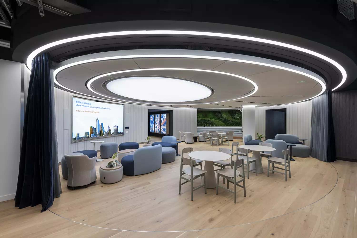 A sleek, modern office lounge area with curved, futuristic ceiling lights creating a central focal point. The space features light wooden flooring, soft pastel seating in shades of blue and grey, and a mix of lounge chairs and round tables with light chairs. On the left, a large screen displays a welcome message alongside a digital panel with changing content. A green living wall adds a natural touch at the back, while dark curtains can be drawn to section off the area. The overall design blends comfort with a clean, high-tech look.