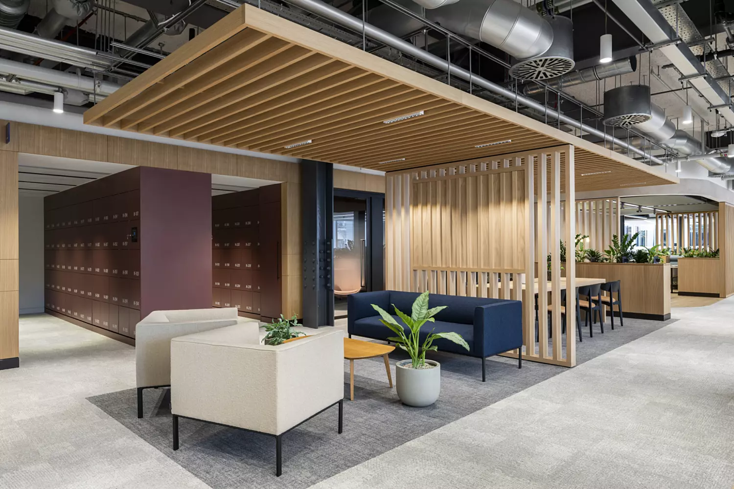 Modern office space with an open layout featuring two beige armchairs, a dark blue sofa and a small wooden coffee table with a potted plant. Behind the seating area is a wooden slatted partition. To the left, maroon lockers line the wall. The ceiling has exposed ducts and pipes and additional workspaces and plants are visible in the background.