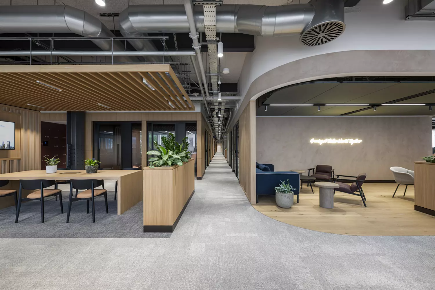 A contemporary office interior with a mix of open and semi-enclosed spaces. On the left, there's a long wooden table with black chairs beneath a slatted timber ceiling feature, surrounded by wooden panelling and indoor plants. A central corridor stretches into the distance, lined with enclosed meeting rooms or offices on either side. On the right, there's a relaxed lounge area with armchairs, a sofa, small round tables and potted plants on light wooden flooring. A softly textured wall at the back features a glowing neon sign that reads “Beautiful things start together.” The ceiling has exposed ductwork, adding an industrial touch.