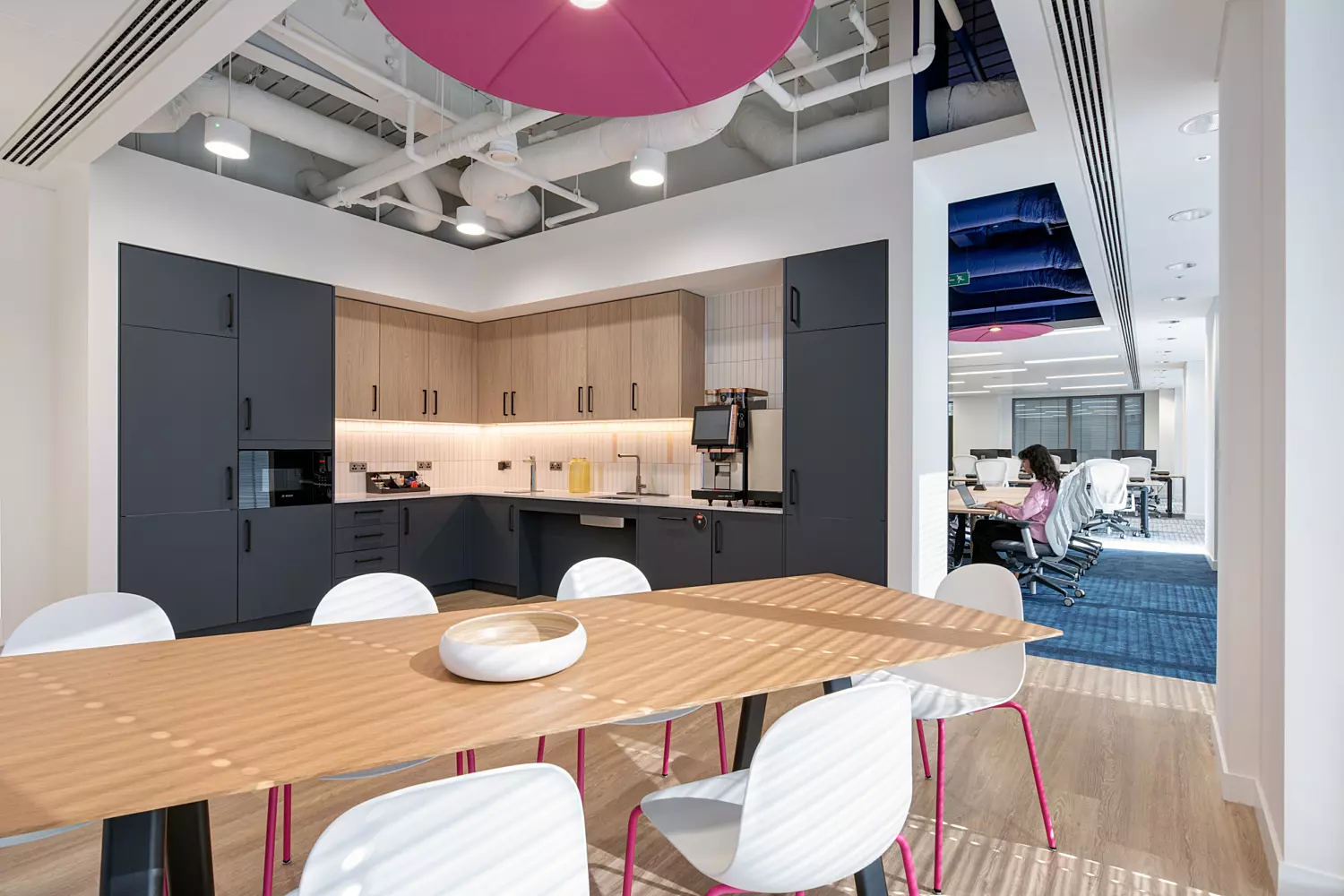 A modern office kitchen and dining area featuring dark cabinetry paired with light countertops, equipped with appliances including a coffee machine. A central wooden dining table is surrounded by white chairs, with a decorative bowl placed on top for a homely touch. The ceiling showcases exposed ductwork and piping, with sections painted in bright colours such as pink and blue, adding a playful industrial flair. In the background, an open doorway reveals an adjoining office space where people are working at desks, contributing to the open-plan layout.