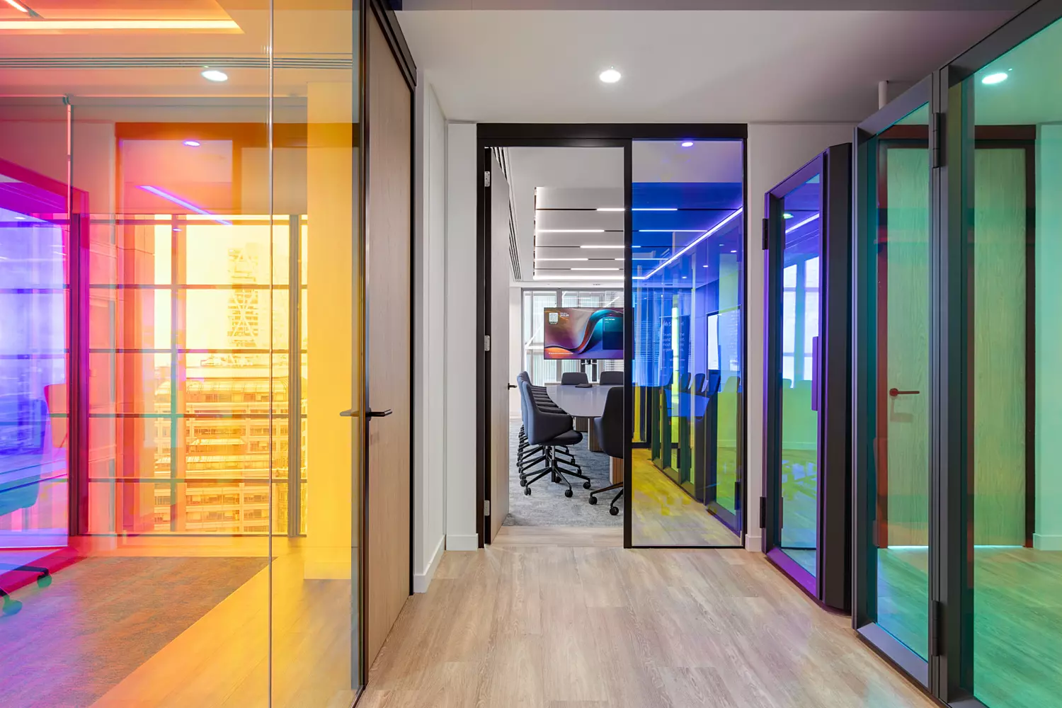 A sleek, modern office hallway lined with glass walls and doors, each tinted in a range of vivid colours including red, yellow, blue, and green. The corridor leads to an open-plan office area, where desks and chairs are visible through the glass at the far end. The flooring is a light-toned wood, contributing to a bright and clean aesthetic, while ceiling-mounted lights provide even illumination throughout the space.