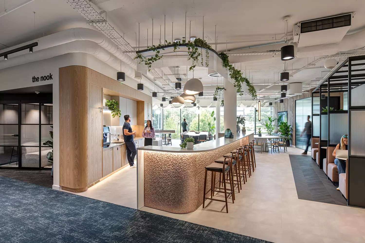 A contemporary office interior showcasing a central curved bar counter with high stools, lit by pendant lights. The bar features a textured façade and is accentuated by ceiling-mounted greenery. To the left, a wooden kitchenette includes a coffee machine and informal gathering space. On the right, semi-private booth seating with frosted glass panels offers quieter work zones. The flooring combines light tiles with dark carpet, while expansive windows provide ample natural light. Exposed ductwork and suspended lighting contribute to the open-plan, industrial aesthetic.
