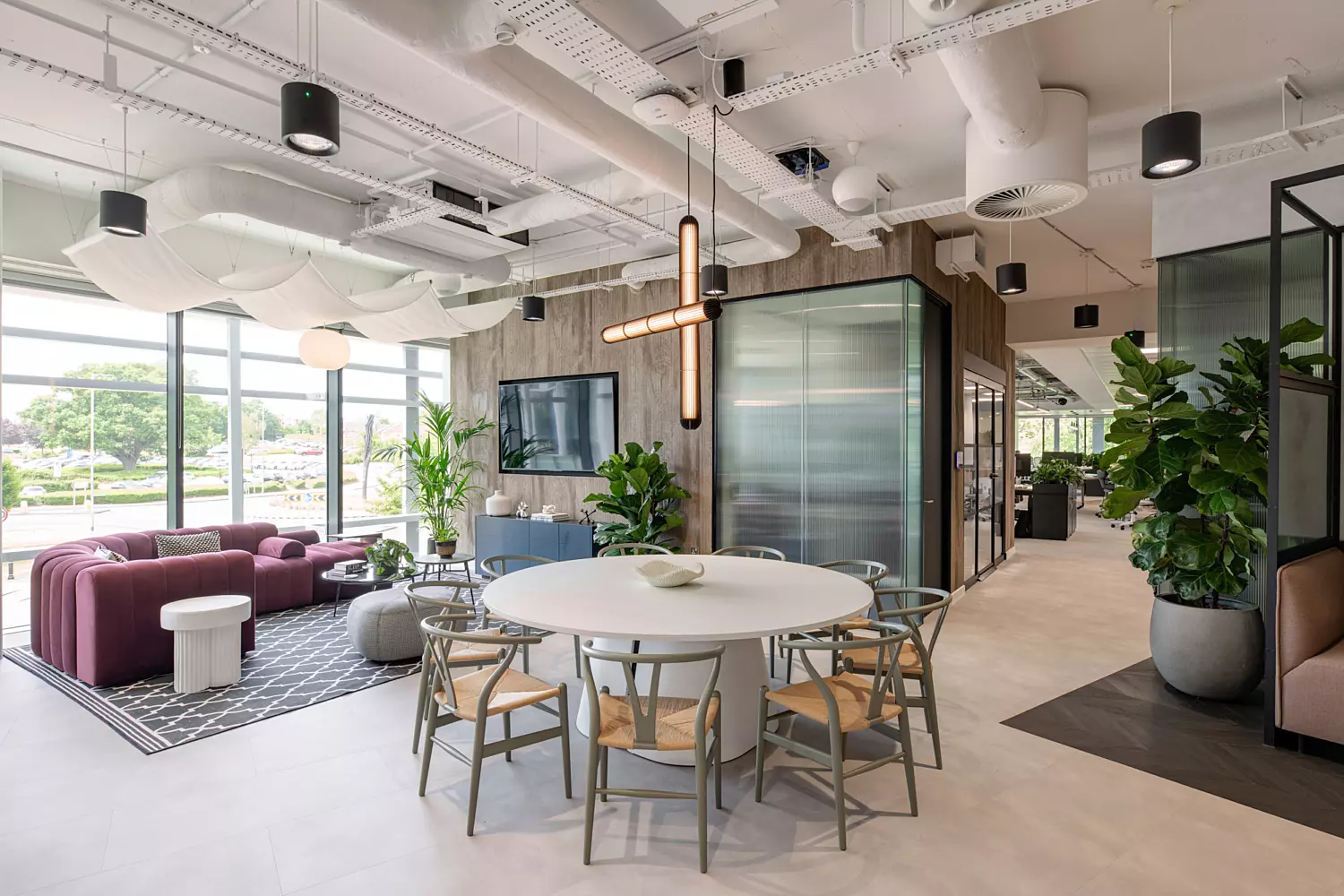 A modern open-plan office featuring a round meeting table with multiple chairs in the foreground. To the left, a lounge area includes a red sofa and white side tables, framed by decorative indoor plants. Large windows flood the space with natural light, while exposed ductwork and piping on the ceiling enhance the industrial design. Contemporary lighting fixtures and enclosed rooms in the background add to the functional and stylish layout.