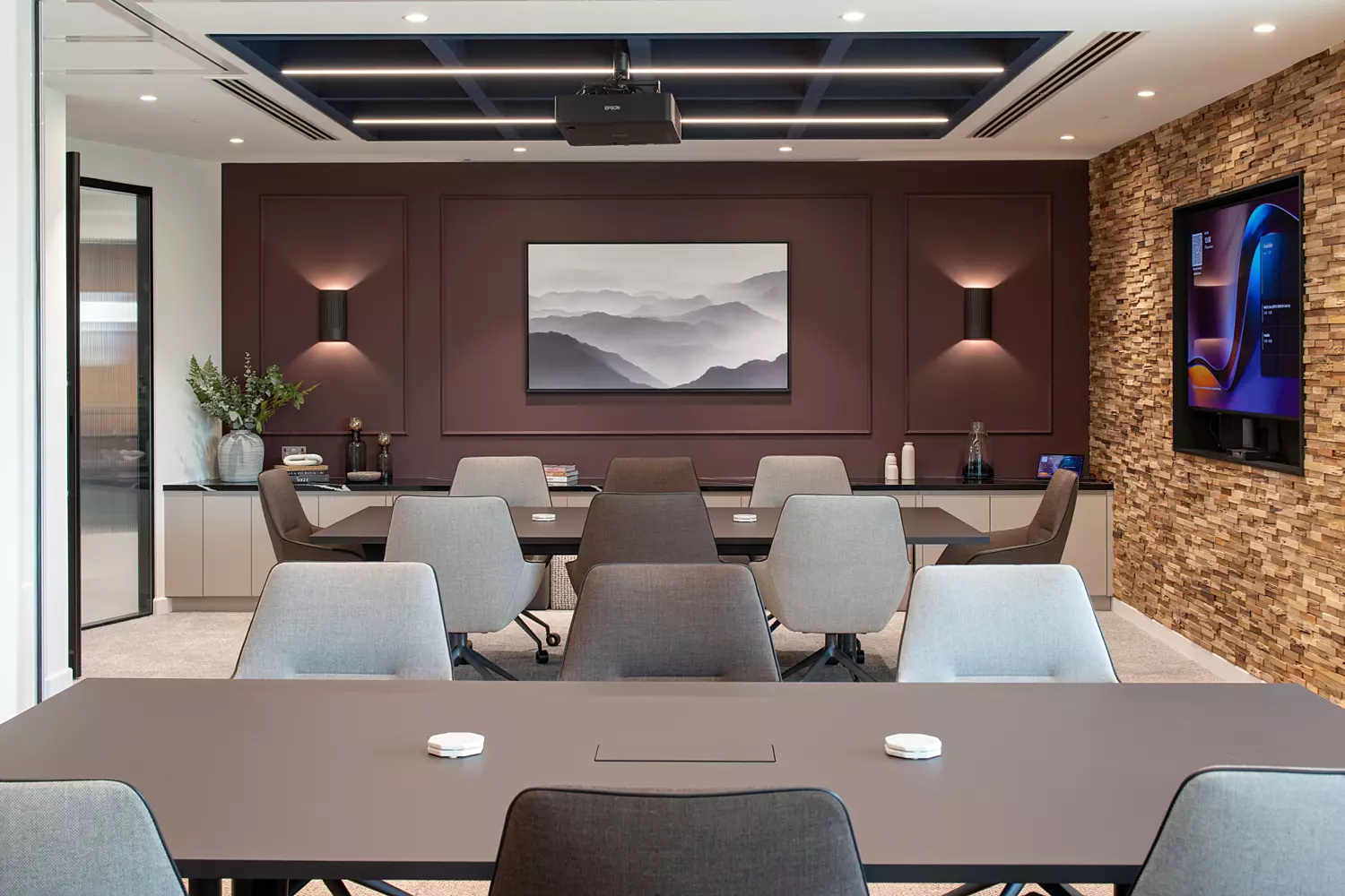 A modern conference room with a large central table surrounded by multiple chairs. A ceiling-mounted projector faces a wall-mounted screen displaying an abstract mountain image. Another screen on the right wall shows additional content. The room features wooden wall panelling with shelves holding plants and decorative items, creating a warm professional atmosphere.