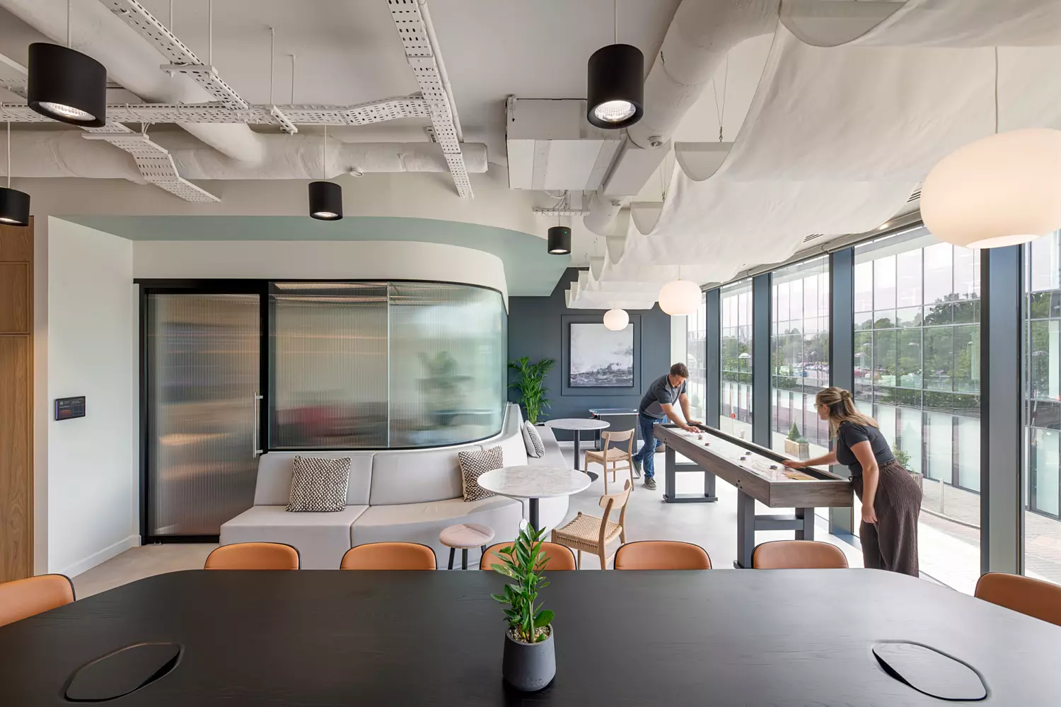A stylish office lounge area with large windows providing ample natural light. The space includes a curved sofa, chairs and stools arranged around tables offering varied seating options. A shuffleboard table is positioned on the right adding a recreational element. The ceiling features exposed ductwork and suspended lighting contributing to the industrial aesthetic. Indoor plants placed on tables and near the back wall enhance the relaxed modern atmosphere.