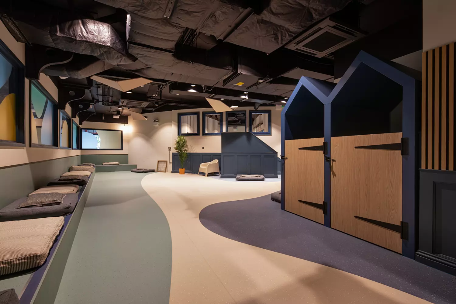 Spacious dog lounge designed with curved flooring in soft blue and cream tones for a playful, flowing effect. Built-in cushioned seating lines one wall, while kennel-style structures with pitched roofs in blue and wood tones provide private spaces. Exposed black ceiling ducts add an industrial edge, complemented by accent panels and integrated lighting.