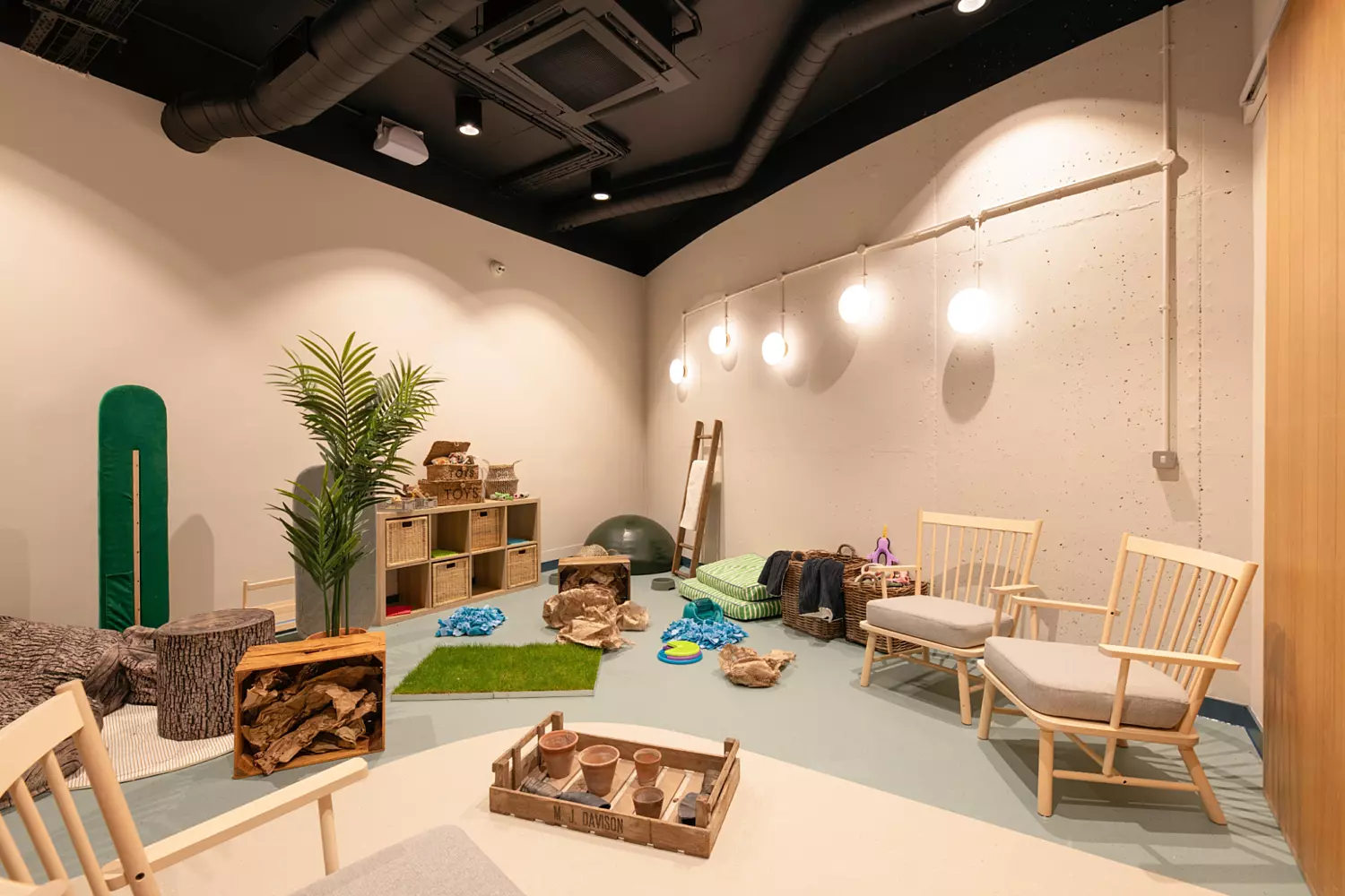 Bright enrichment room with a playful, natural theme. Light walls and soft flooring in green and beige tones set a calming backdrop. The space includes wooden shelving filled with textured play items, faux grass mats, and natural elements like logs and stones. Pendant globe lights and simple wooden chairs complete the relaxed, creative environment.