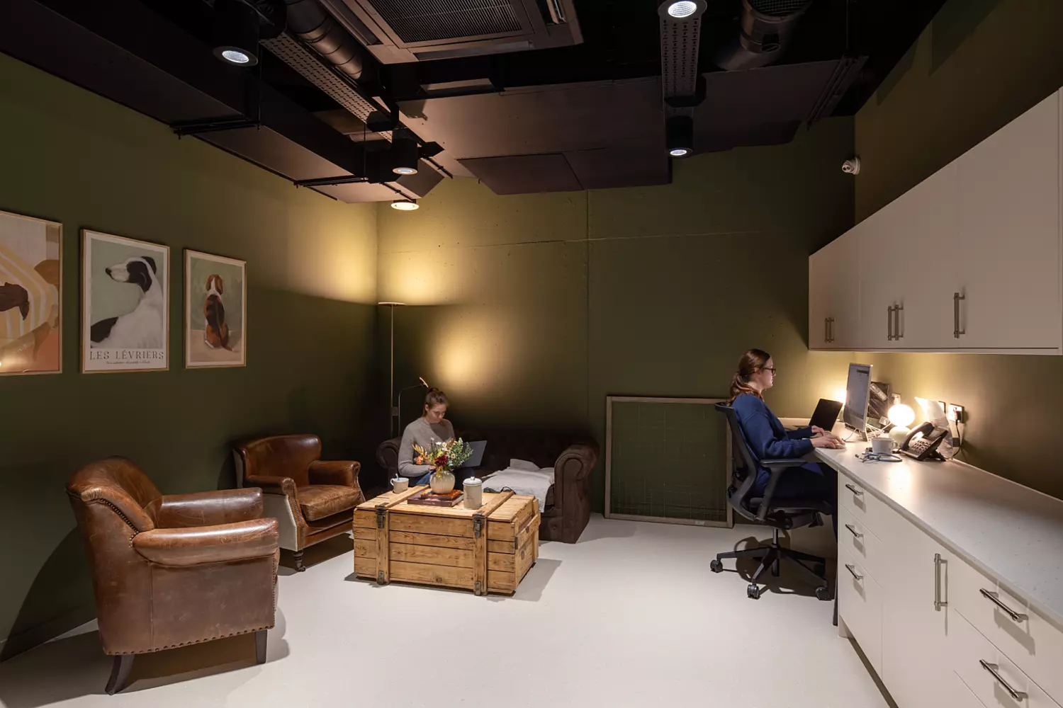 Cosy staff office with deep green walls and contrasting white cabinetry. The space combines functionality with comfort, featuring leather armchairs, a rustic wooden coffee table, and framed dog-themed artwork. Overhead black ceiling panels and spotlights create a modern, slightly industrial aesthetic.