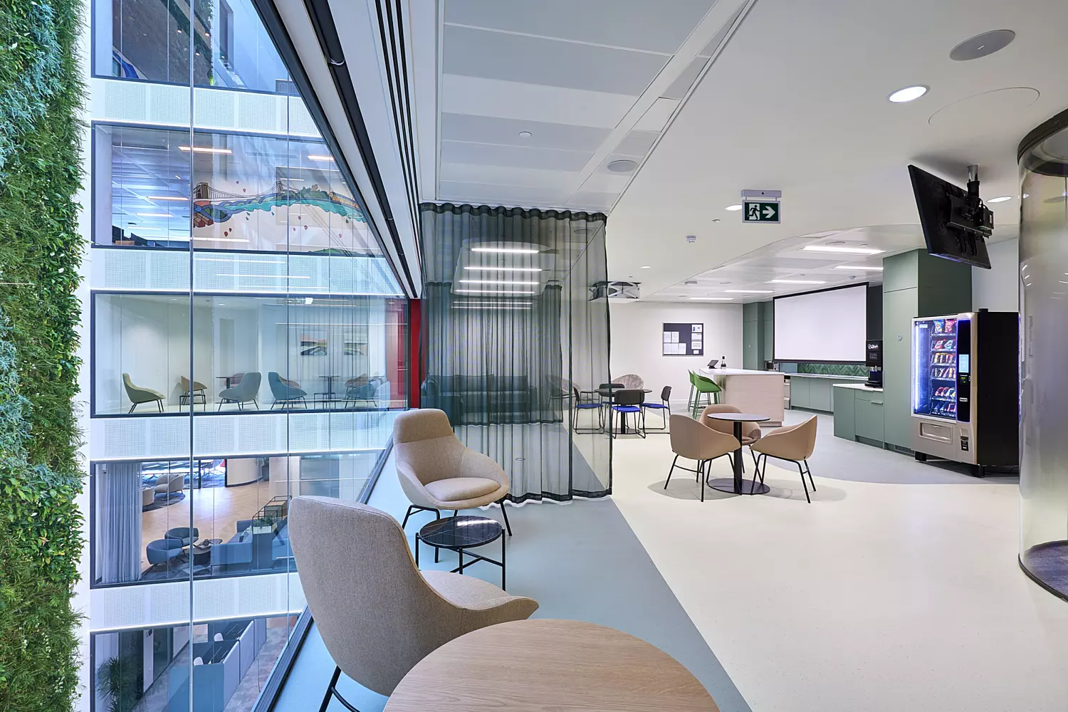 Modern breakout space with floor-to-ceiling glass overlooking an open atrium. The area features soft seating in neutral tones, round tables, and a vending zone with a green accent wall. A striking vertical living wall adds biophilic design, while sheer black curtains create flexible zoning. The space is bright and open, with clean lines and contemporary finishes that encourage informal collaboration.