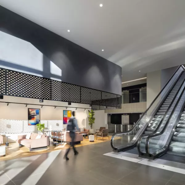 A modern indoor office space featuring a relaxed seating area with sofas, chairs and tables, accented by colourful wall art. An escalator on the right leads to an upper level, surrounded by greenery. Large windows fill the space with natural light, creating a bright, welcoming atmosphere.