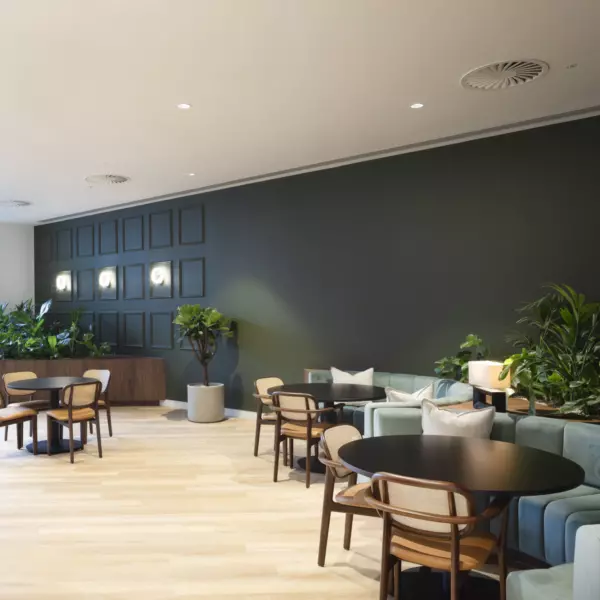 A modern lounge area with wooden flooring and varied seating including round tables with chairs and a long cushioned sofa. Green plants in pots and built-in planters add natural texture. The back wall features dark square panelling with integrated lighting, while large windows on the left bring in natural light.
