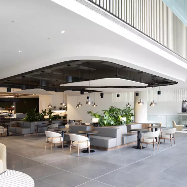 A spacious modern indoor area with sofas and chairs arranged around tables, illuminated by natural light from large windows. Hanging ceiling lights and greenery enhance the relaxed atmosphere. The tiled grey flooring leads to a café-style counter in the background, with additional seating visible further into the space.