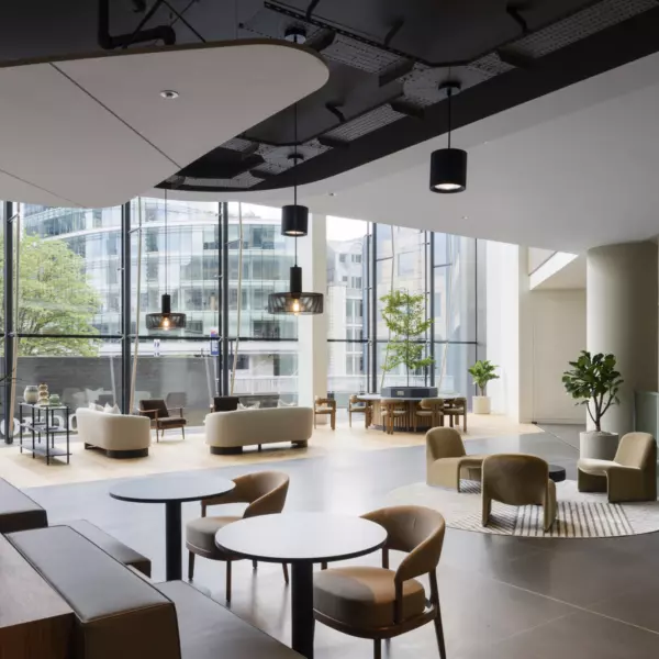 A spacious modern lounge area with floor-to-ceiling windows offering city views. Seating includes round tables with chairs, sofas and armchairs arranged for comfort and flexibility. Hanging light fixtures and ceiling panels add visual interest, while potted plants bring greenery to the contemporary, welcoming interior.